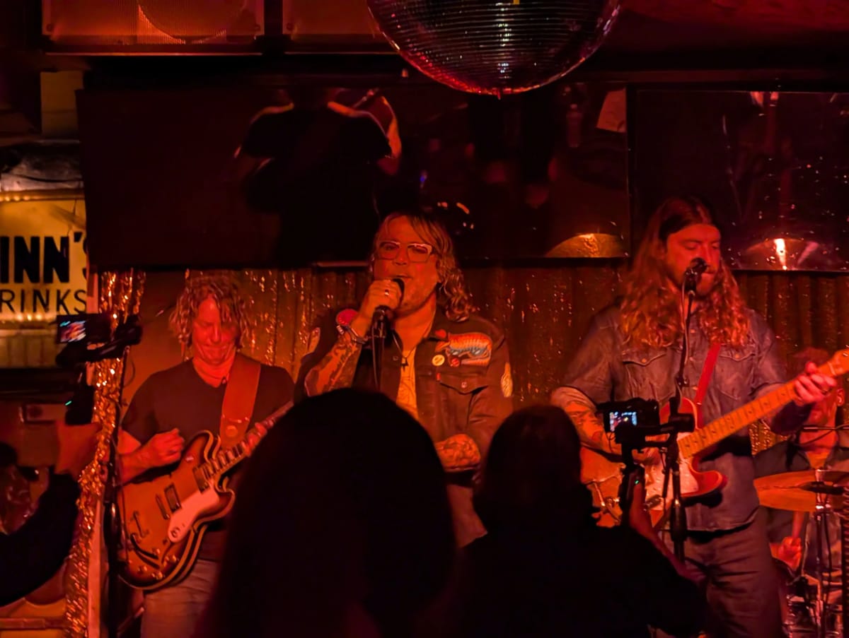 Three performers sing and play guitars on a red-lit club stage beneath a mirror ball.